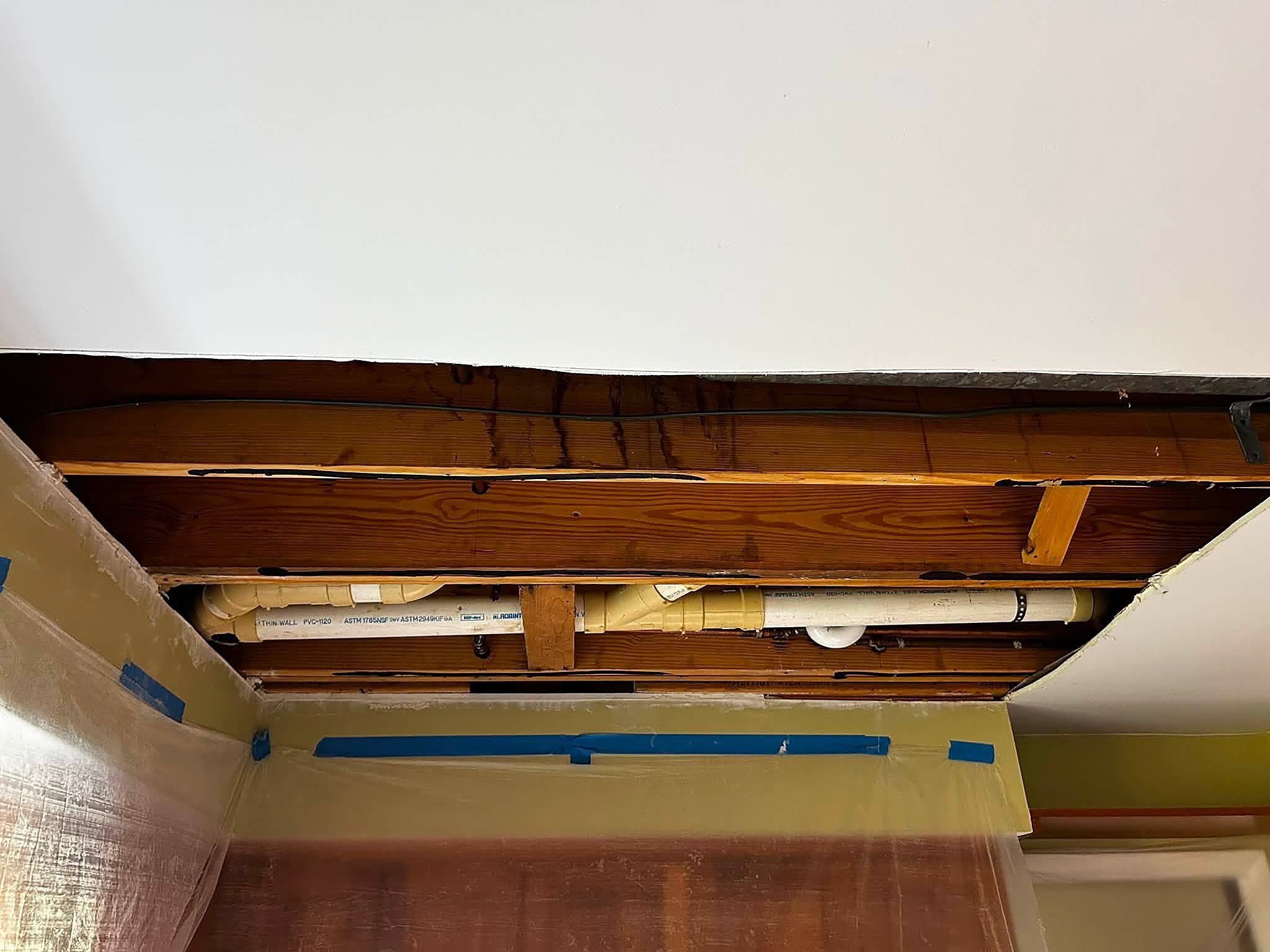 Opened ceiling cavity showing drain pipes and water-stained joists above a living space