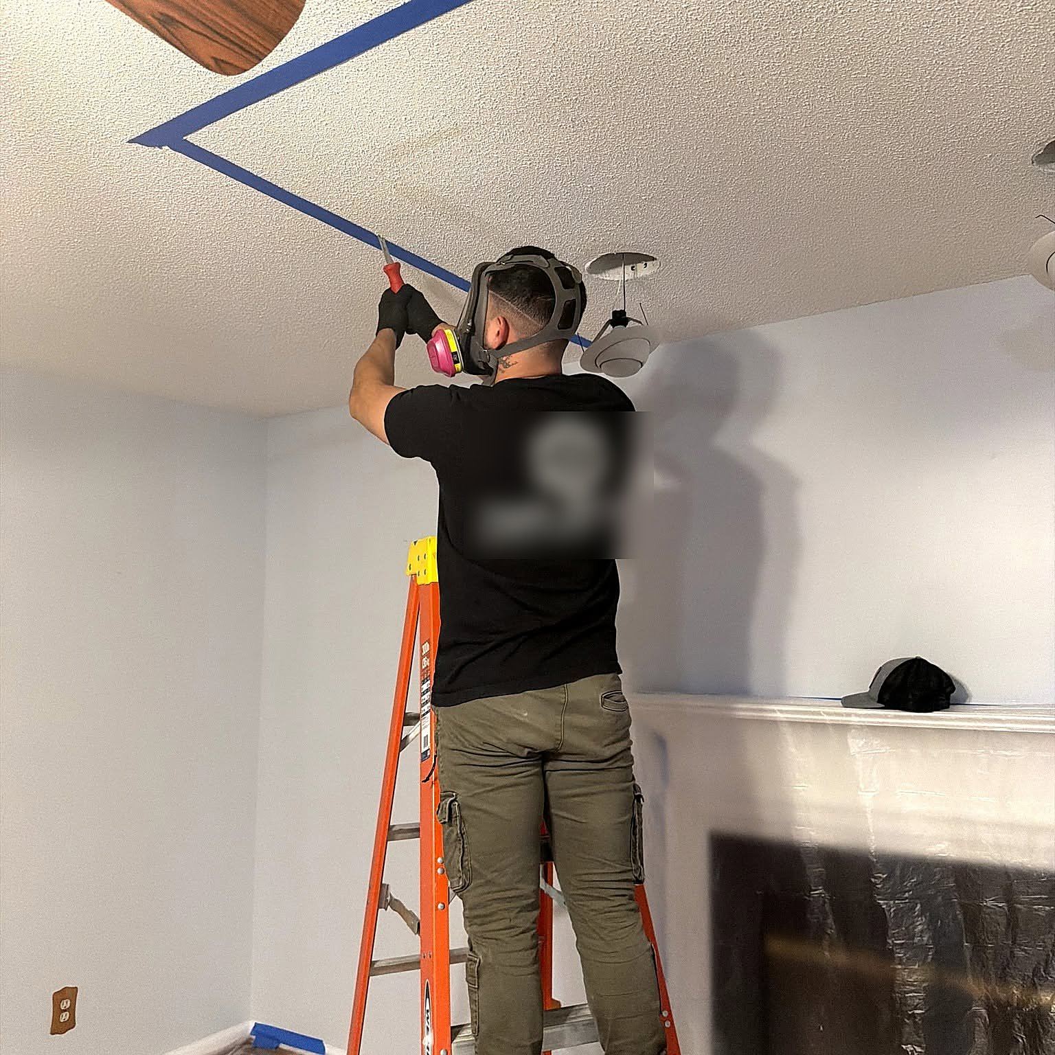 Technician in a respirator working on a textured ceiling with painter tape marking the repair area