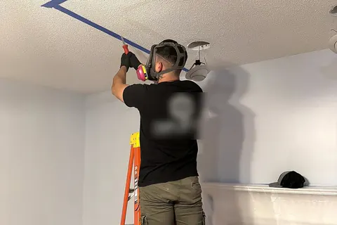 Technician in a respirator working on a textured ceiling with painter tape marking the repair area